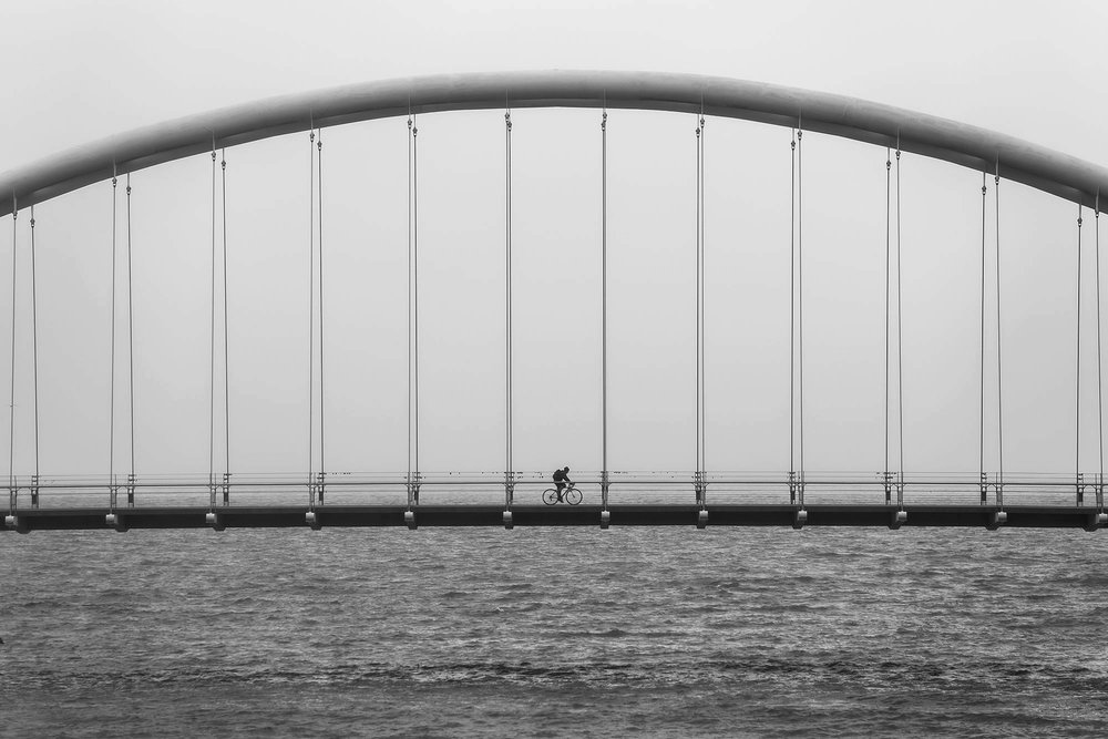 black and white photo of cyclist on suspension bridge over water