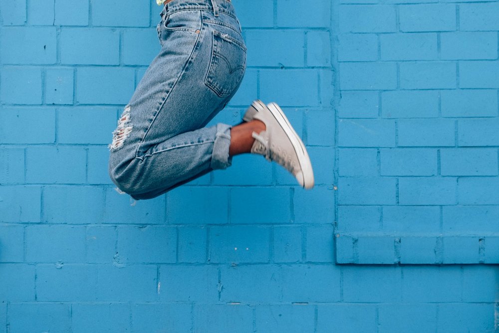 legs of a person in jeans and trainers jumping in front of a blue brick wall